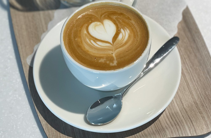 Das Foto zeigt einen Kaffee in einer weißen Tassen auf einem weißen Untersetzer mit einem Löffel